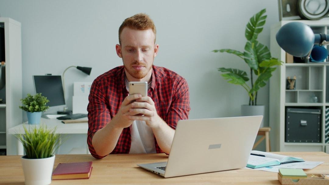 Man with red hair using smartphone at desk with laptop.